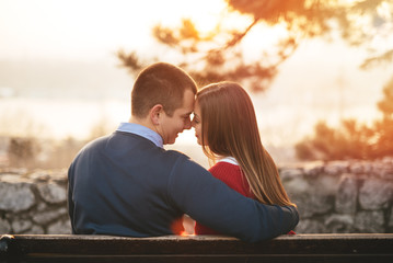 Young couple sitting on park bench and enjoying in love. River, city and beautiful sunset colors in background.