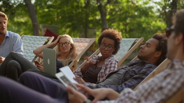 Five interracial friends lying on recliners in park with laptop notebooks tablets speaking smling chatting in slowmotion