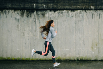 Female urban athlete running outside against a concrete wall. Sporty woman training and exercising.