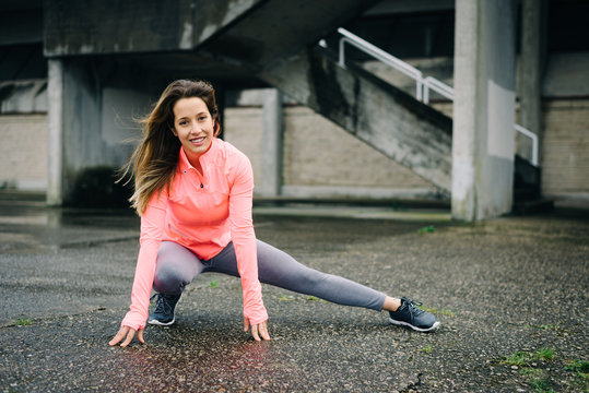 Sporty young woman stretching long adductor for warming up before urban fitness workout or running training. Caucasian female athlete doing leg stretch exercise outside on rainy winter day.