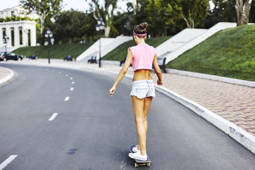 Beautiful hipster girl in a jeans shorts and pink shirt walking with skateboard near the green park, sexy style, adult, long legs, brunette, white shoes, sexy model, posing on camera