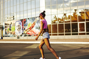 Obraz premium Beautiful hipster girl in a jeans shorts and pink shirt walking with skateboard near the green park, sexy style, adult, long legs, brunette, white shoes, sexy model, posing on camera