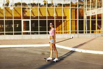 Obraz premium Beautiful hipster girl in a jeans shorts and pink shirt walking with skateboard near the green park, sexy style, adult, long legs, brunette, white shoes, sexy model, posing on camera