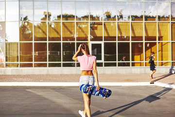 Beautiful hipster girl in a jeans shorts and pink shirt walking with skateboard near the green park, sexy style, adult, long legs, brunette, white shoes, sexy model, posing on camera