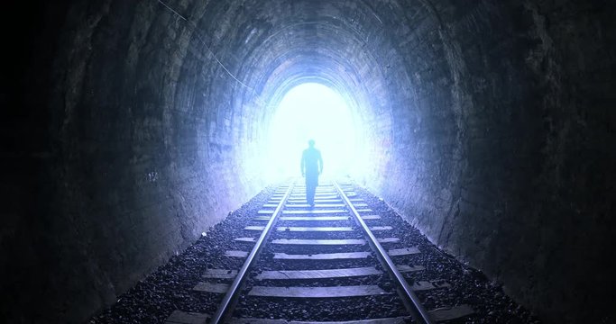 Man goes by railway tracks through dark tunnel toward bright light in the end