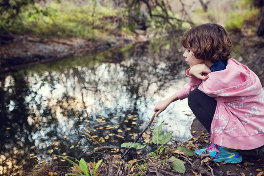 Side view of girl holding stick while sitting by stream in forest