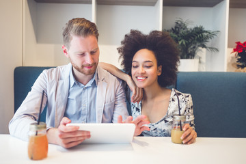 Happy middle age interracial couple sitting in cafe bar, smiling and looking at white tablet.