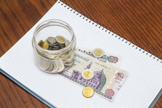 Glass Full of Coins on Banknotes with Notebook on Wooden Desk