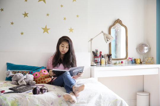 Teenage girl sitting on bed and reading book at home
