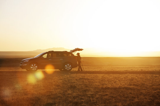 Side View Of Hiker Standing By Car On Field Against Clear Sky During Sunset