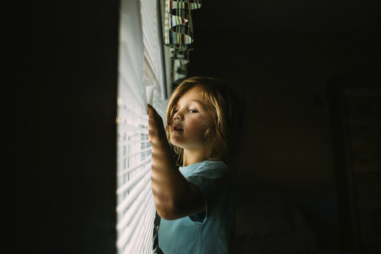 Girl looking though window blinds while standing in darkroom at home