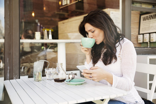 Woman Drinking Coffee While Using Smart Phone At Sidewalk Cafe