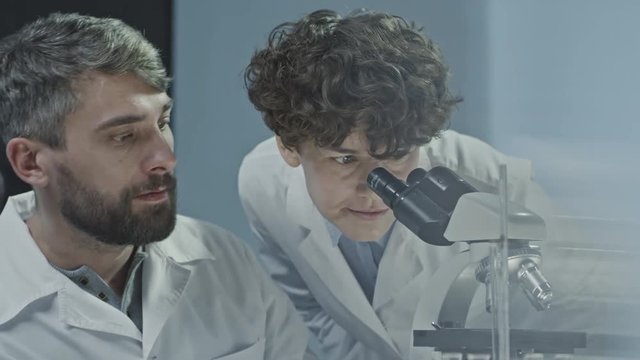 Male scientist with beard observing sample and asking opinion of his female colleague taking off glasses and looking into microscope 