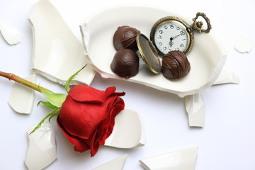 Shattered bowl with a rose and chocolates next to a pocket watch.