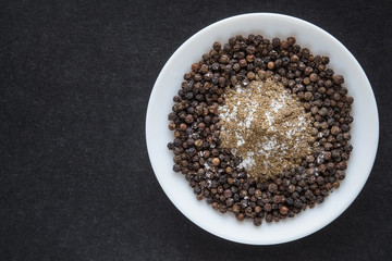 Black pepper grains with ground pepper and a little bit salt in the white dish on the dark background.