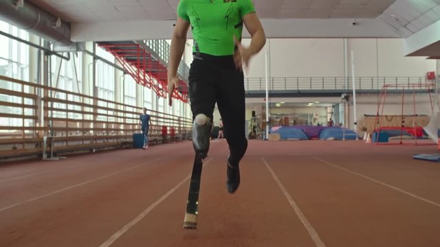 Dolly shot of amputee athlete with prosthetic leg running in slow motion at indoor stadium 