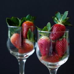 Square photo of two vine glasses with fresh red strawberry with green leaves on black background