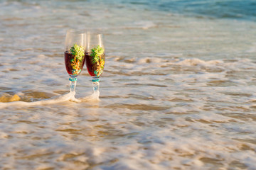 Glasses of champagne on the beach against blue sea. Pair of wine glasses on the beach. two glasses in wave of the sea on the beach. Valentine's Day on island, romantic evening, 


