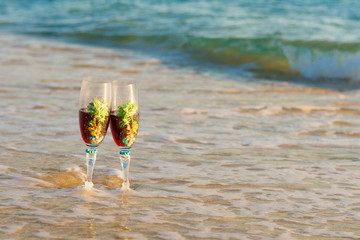 Glasses of champagne on the beach against blue sea. Pair of wine glasses on the beach. two glasses in wave of the sea on the beach. Valentine's Day on island, romantic evening, 

