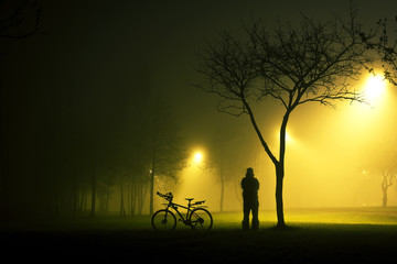One man is standing and holding the bicycle in foggy and mysterious park at night