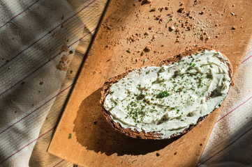 Slice of grilled bread with burnt black edges, cream cheese and green basil. Bread is on wooden board and striped tablecloth. Lots of crumbs around in the sun golden light with shadows and reflection.