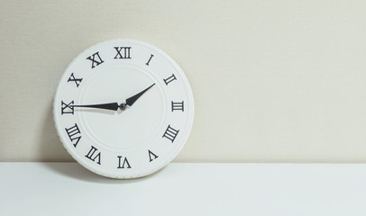 Closeup white clock for decorate show a quarter to two p.m. or 1:45 p.m. on white wood desk and cream wallpaper textured background with copy space
