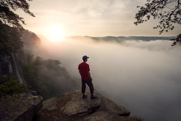 A white man wearing a red shirt looks out over the New River Gorge in West Virginia on a misty morning at sunrise.