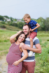 Fototapeta premium Pregnant family of three in expectation of baby. Father mother and son playing together in day outside. Happy people hugging. Smiling laughing baby sitting on shoulders of daddy.