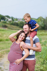 Fototapeta premium Pregnant family of three in expectation of baby. Father mother and son playing together in day outside. Happy people hugging. Smiling laughing baby sitting on shoulders of daddy.