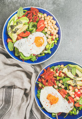 Healthy breakfast bowls with fried egg, chickpea sprouts, seeds, fresh vegetables and greens over grey concrete background, top view. Clean eating, dieting, healthy lifestyle, vegetarian food concept