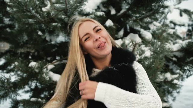 Funny woman fools around in pine winter snowy forest outdoors. Attractive Russian female in white woolen sweater, black fur vest walks, has fun, makes mustache from long fair straight hair. Cute lady