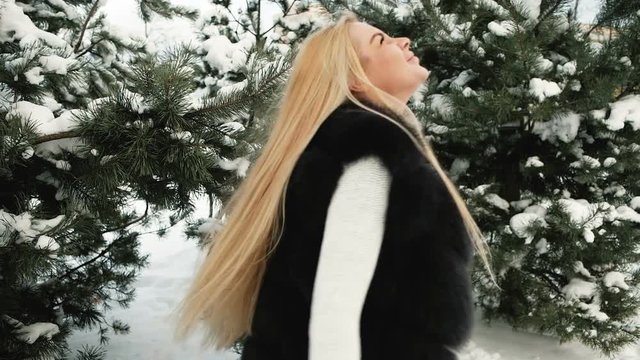 Fantastic blonde is turned against background of snow covered pines. Wonderful girl is dressed in white knitted sweater, black fur vest and pearl woolen mittens. She blinks with happiness and joyfully