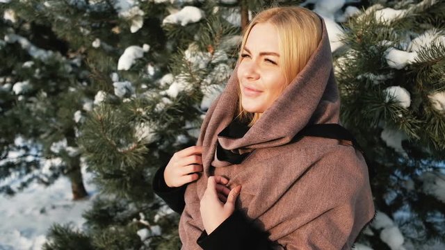 Dreaming blond woman looks far away in winter pine forest outside. Lovely Russian beautiful female in warm clothes enjoying good frosty weather in sunny day. Attractive charming blonde in black coat