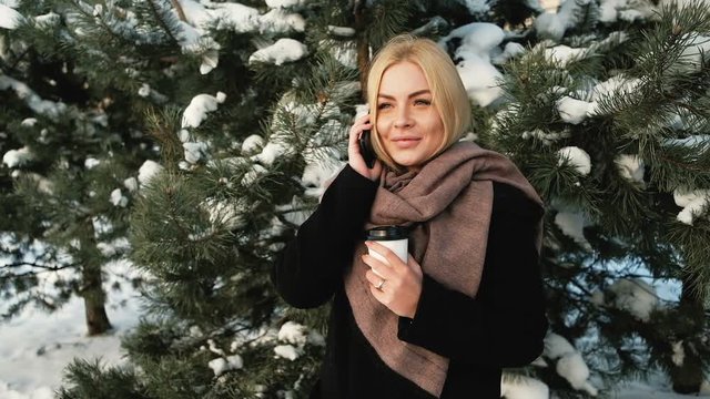 A woman with a glass in forest talking on the phone and smiling. She stands at the pine tree, and carrying on a conversation. European woman dressed in a black coat, a brown scarf. A pleasant