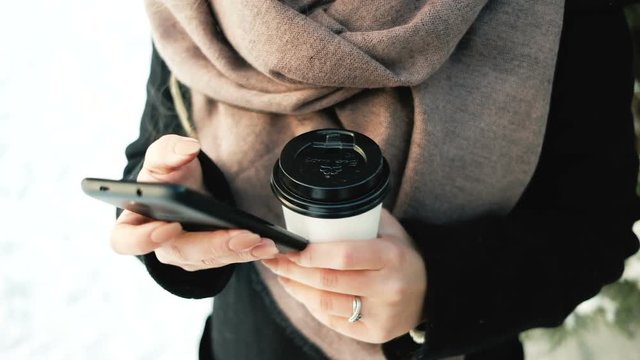 Young female with phone, thermos cup outdoors in winter. Close up hands of Walking lady looking, touches Smartphone in pine forest. Attractive Russian woman in black warm coat, beige scarf walks, has