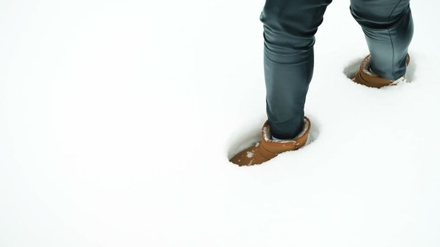 Female legs in brown uggs leave traces in the white pure snow. Slim woman wearing black leather skin-tight leggins and big flat-heeled boots with a belt puts one foot in front of the other. She is