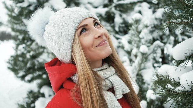 Young pretty woman warm in ray of sun in winter forest. We see attractive slim lady with long beautiful blond hair in red Down jacket with gold zip, hood, woolen grey scarf and cap with small jewelry