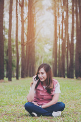 A beautiful asian woman talking on the smart phone with feeling relax and smiley face , sitting in pine tree garden , green nature background
