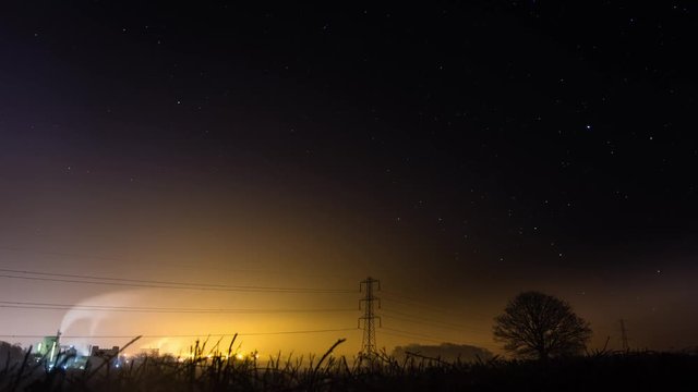 Factory and trees in front of revolving night sky timelapse. Industrial landscape in Wiltshire, UK, with moving stars, chimneys and electricity pylons. 