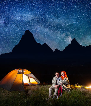 Sweety couple tourists covered with a plaid sitting together near campfire and shines tent at night. High mountains, starry sky, Milky way on the background