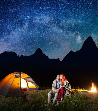 Happy couple tourists covered with a plaid sitting together near campfire and shines camp at night under stars and looking to the starry sky and Milky way. Silhouette of high mountains on background