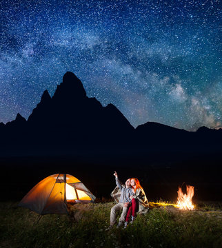 Tourist man showing red-haired woman at the stars in the sky. Couple sitting near the camp on the background silhouette of the high mountains at night
