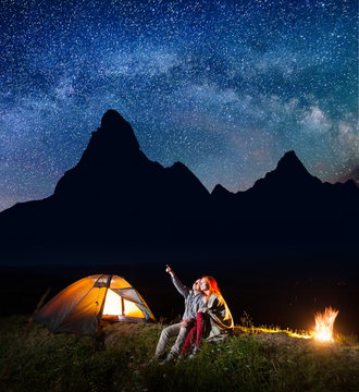 Male backpacker showing his girl at the stars and Milky way in the night sky. Happy couple covered with plaid sitting near the glowing tent and campfire. On the background silhouette of the mountains