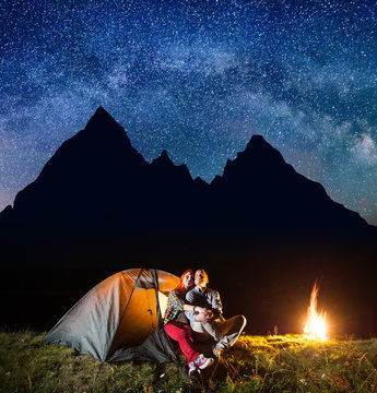 Two hikers having a rest in his camp at night near campfire under shines starry sky on the background silhouette of the mountains. Astrophotography