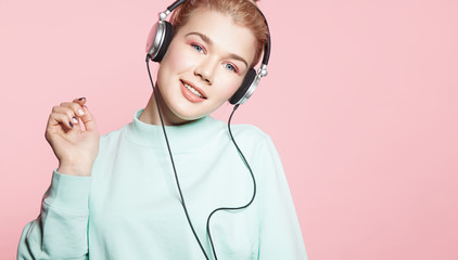 Beautiful woman in headphones listening to music smiling with closed eyes standing on a pink background in a blue sweatshirt