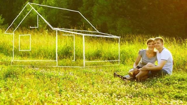 Young happy couple sitting on green grass in park and dreaming about they own house. Mortgage concept.