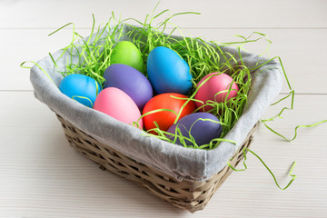Easter wicker basket with colored eggs on white wooden board.