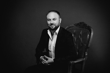 portrait of young man in black suit sitting on luxury chair on dark background monochrome