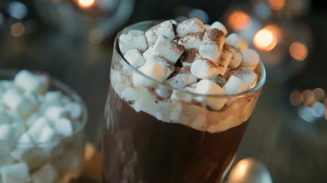 Beautiful composition - hot chocolate with marmalade and chocolate pieces in a transparent glass. The glass stands on a wooden stand. Behind the candles are burning. Close up