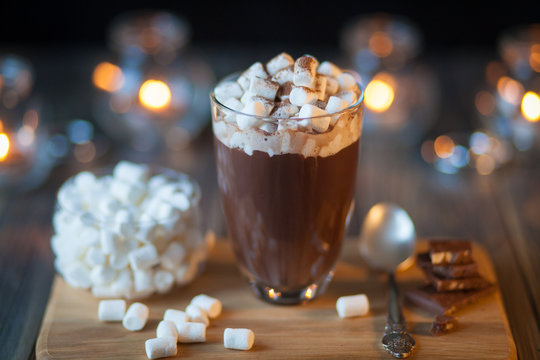 Beautiful Composition - Hot Chocolate With Marmalade And  Pieces In A Transparent Glass. The  Stands On  Wooden Stand. Behind The Candles Are Burning